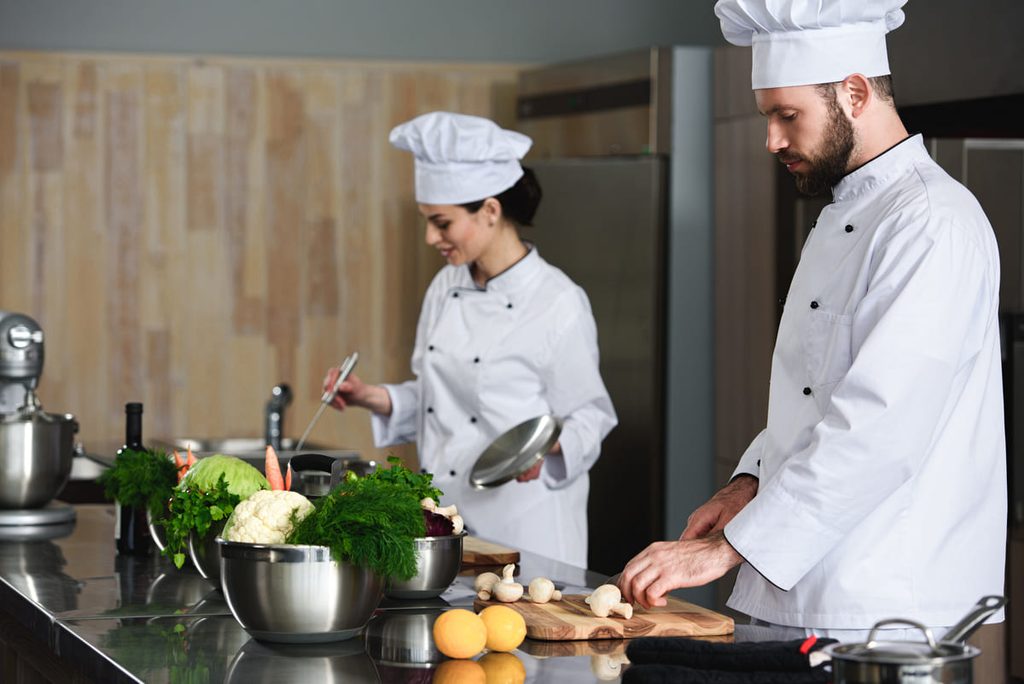Chef y ayudante de cocina preparando y cortando ingredientes en una cocina profesional.